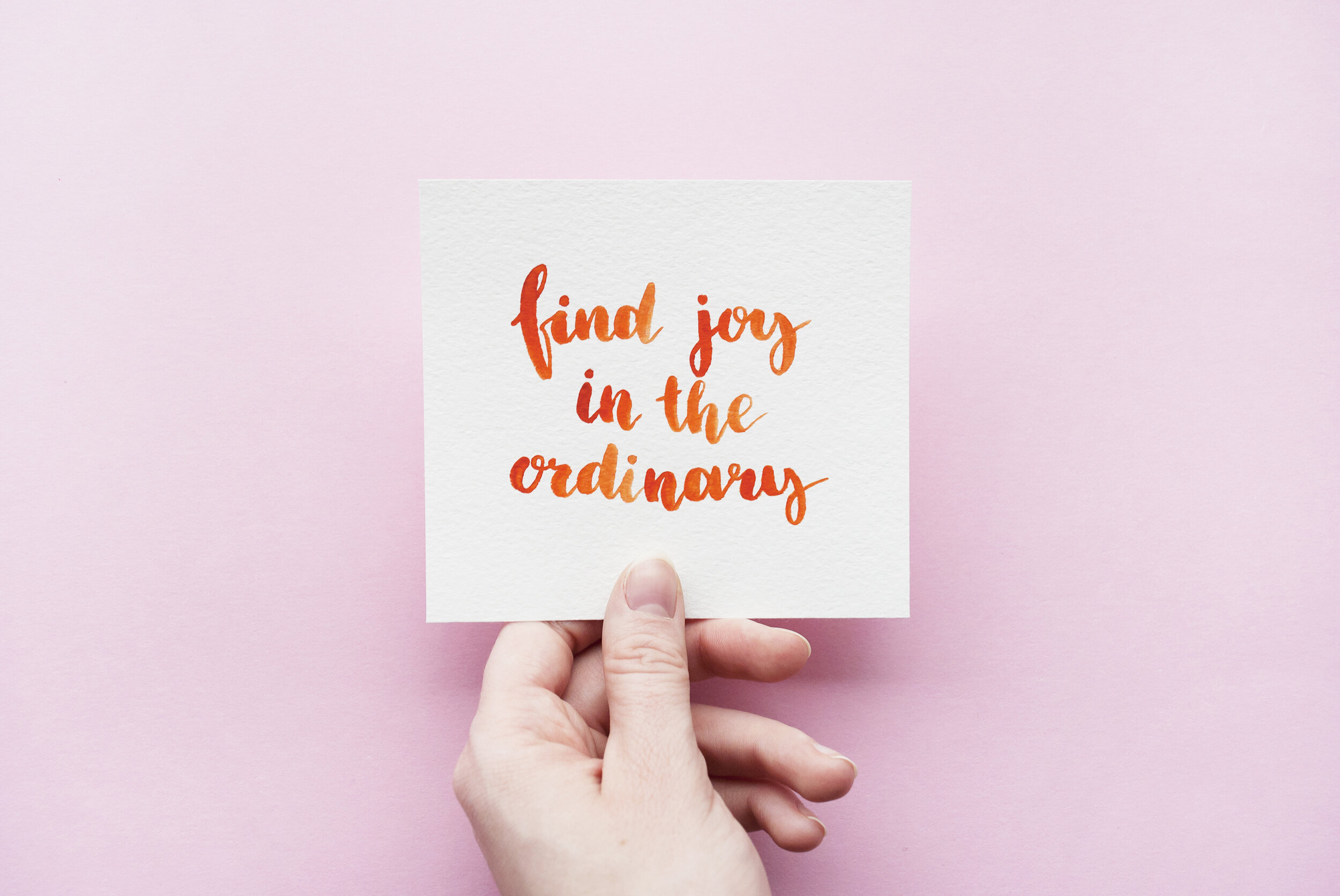 Minimal composition with girl's hand holding card with inspirational quote "Find joy in the ordinary" written in calligraphy style on paper on a pink background. Flat lay (Copy)